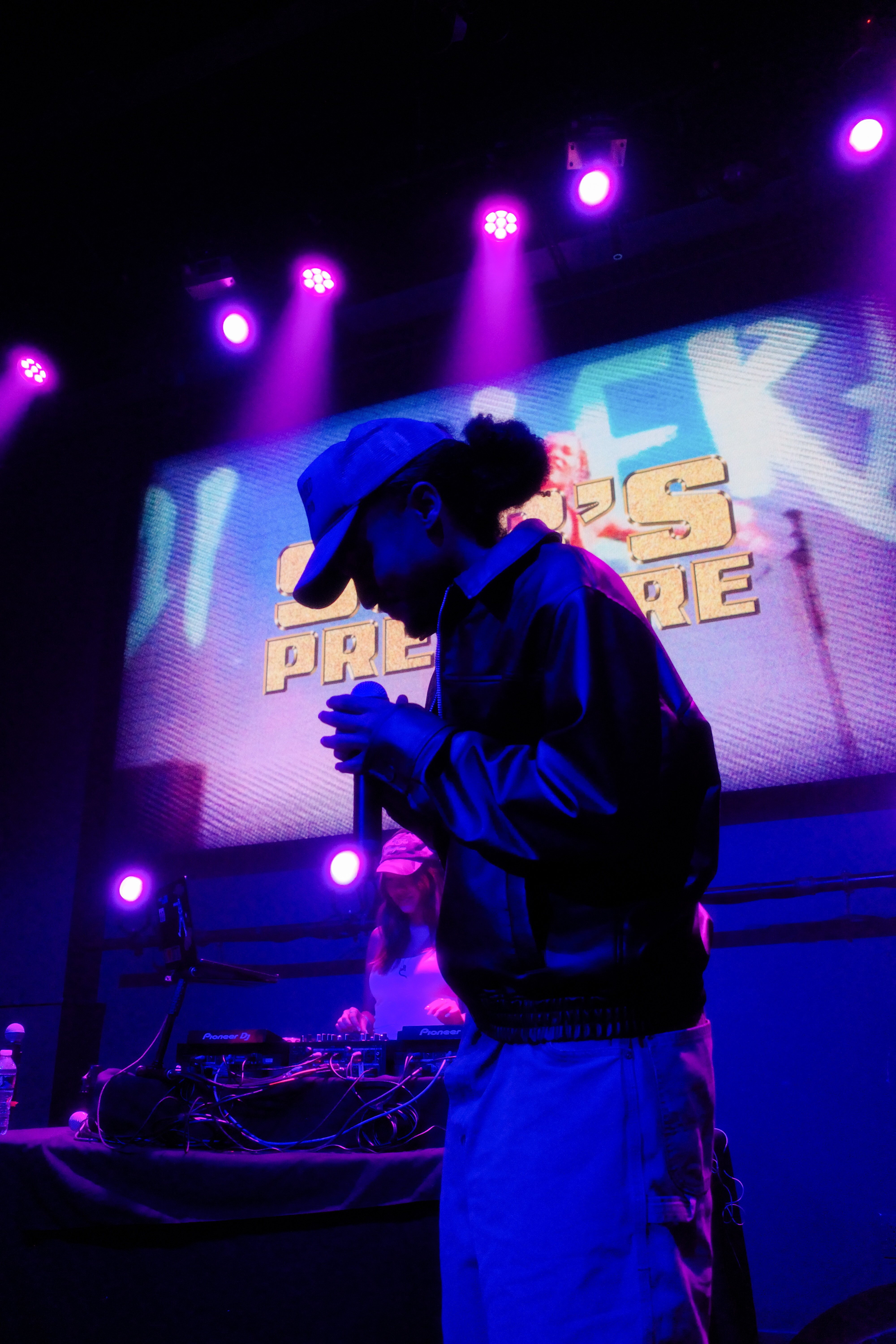 Kuhsigh performing under blue and purple stage lighting, holding microphone, looking down